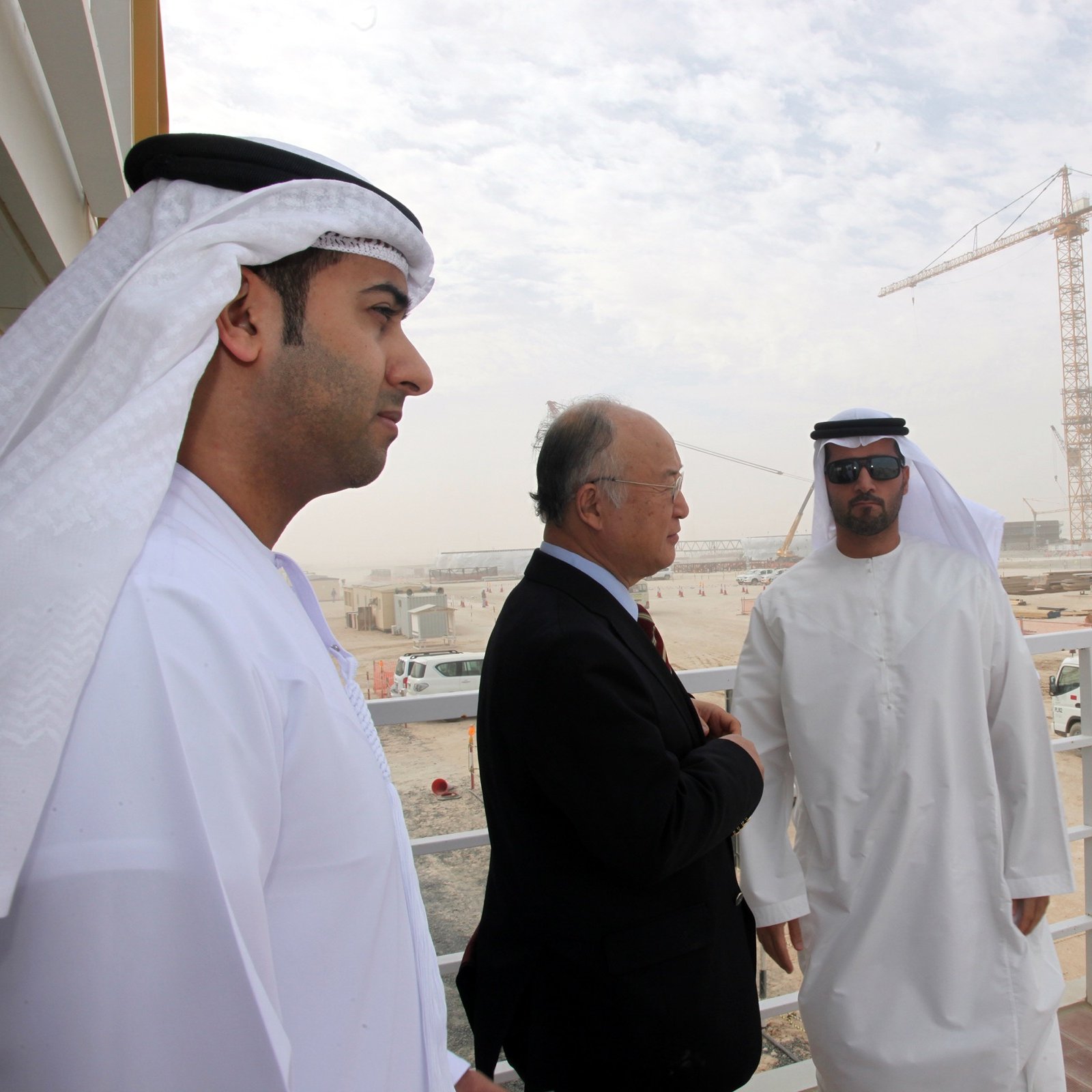 L'image montre trois hommes, apparemment en discussion, sur un balcon ou une plateforme. Ils sont vêtus de vêtements traditionnels des Émirats, comprenant des kandouras et des ghutras. En arrière-plan, on peut voir un site de construction avec des grues et des véhicules, suggérant un projet de développement en cours. L'environnement est poussiéreux et légèrement brumeux, typique des zones de construction dans des climats chauds. Les hommes semblent sérieux et engagés dans leur conversation.