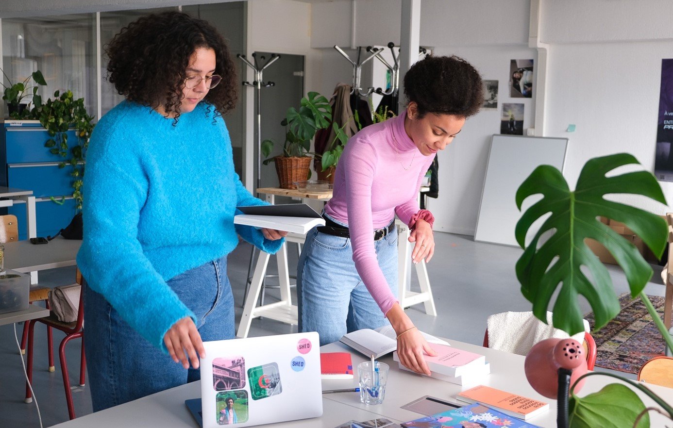 L'image montre deux femmes dans un environnement de travail créatif. L'une d'elles, vêtue d'un pull bleu, semble arranger des choses sur une table. L'autre femme, en pull rose, est en train de manipuler des livres ou des documents. Sur la table, il y a un ordinateur portable, des livres et quelques accessoires de bureau. L'espace est lumineux et décoré de plantes, ce qui contribue à une ambiance agréable et inspirante.