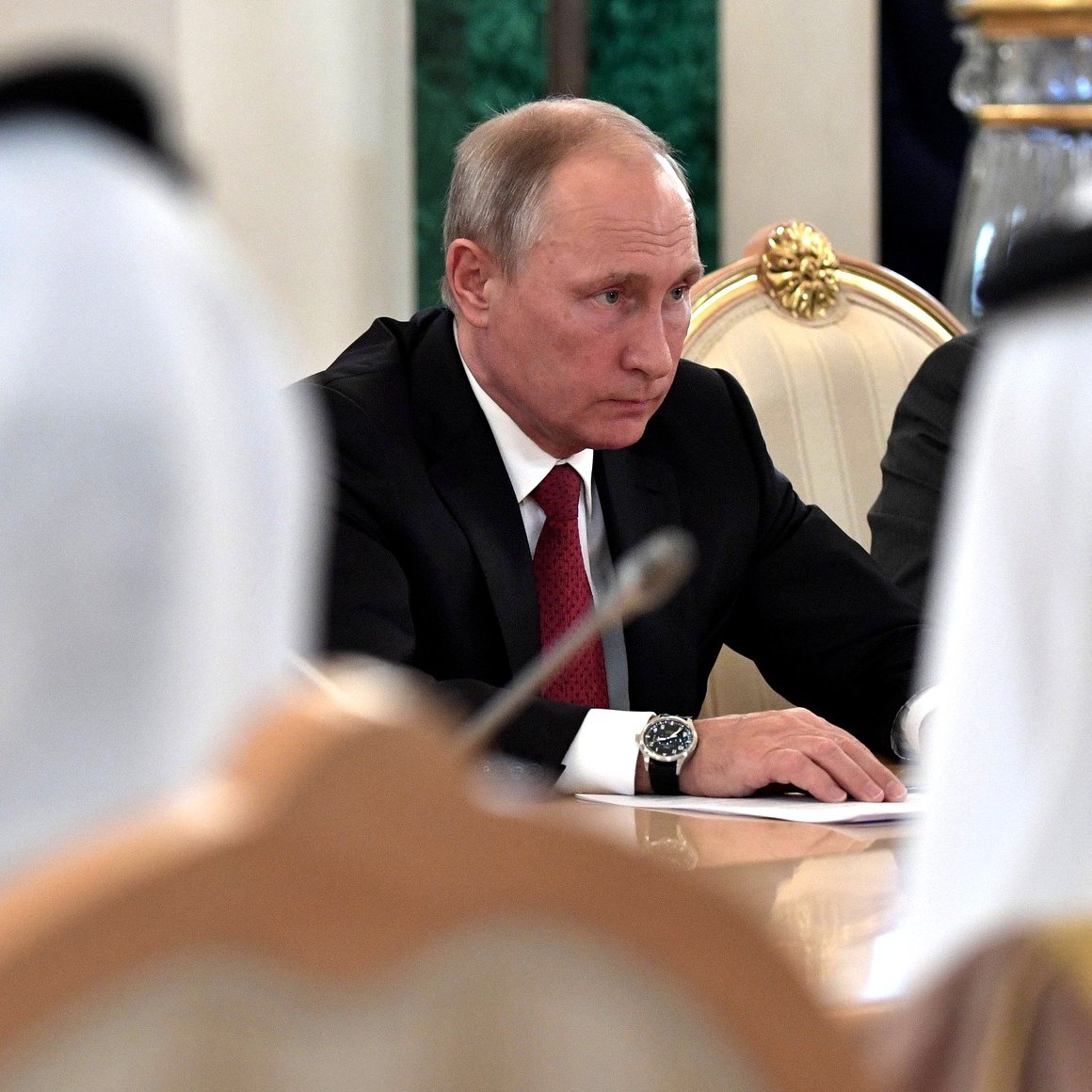 در این تصویر، فردی که به نظر می‌رسد ولادیمیر پوتین است، در حال نشستن در یک جلسه به سمت جلو نگاه می‌کند. او کت و شلوار مشکی به همراه یک رابعه قرمز به تن دارد. در اطراف او، چهره‌هایی از دیگر شرکت‌کنندگان در جلسه دیده می‌شود، اما جزئیات آن‌ها قابل تشخیص نیست. فضای عمومی جلسه جدی به نظر می‌رسد و میز مستطیلی به همراه دکوراتورهای رسمی دیده می‌شود.