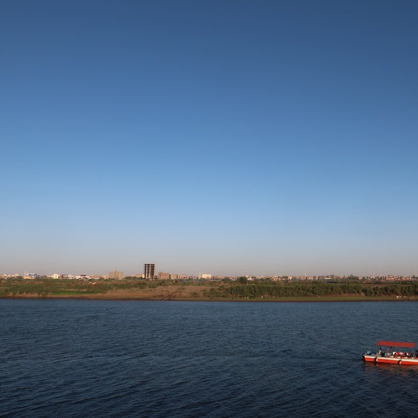 تظهر في الصورة منظرًا طبيعيًا جميلًا يتضمن نهرًا هادئًا ينساب تحت سماء صافية وزرقاء. على جانب النهر، يمكن رؤية مجموعة من الأشجار والنباتات التي تزين الساحل. بينما في وسط النهر، تبحر قارب صغير ذو سقف برتقالي، حيث يبدو أن هناك أشخاصًا يستمتعون بالرحلة. في الخلفية، تمتد المدينة مع المباني السكنية، مما يضفي طابعًا حضريًا على المشهد الطبيعي.