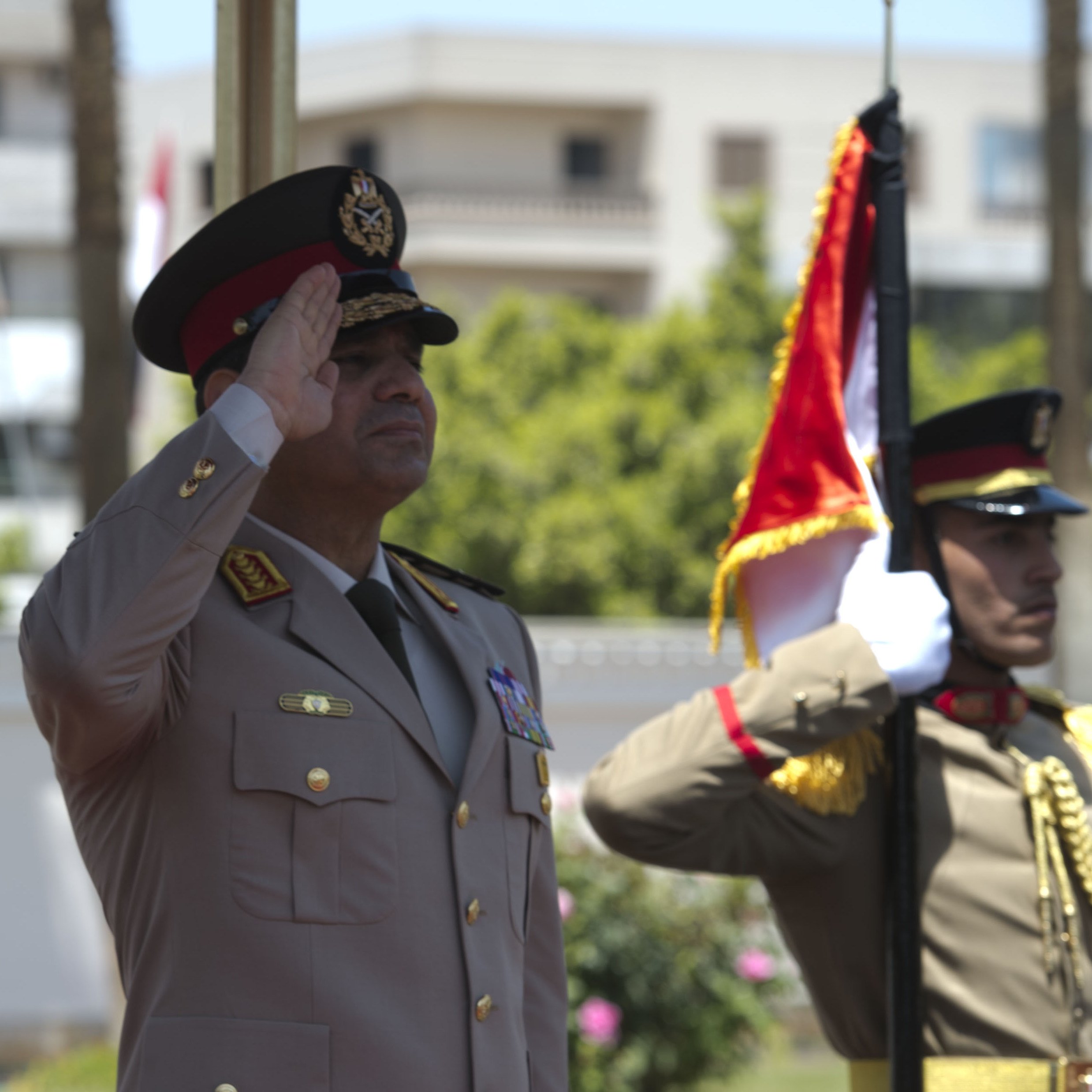 L'image représente deux officiers militaires en uniforme. L'un d'eux, probablement un haut responsable, effectue un salut militaire, tandis que l'autre maintient un drapeau. L'environnement semble être une cérémonie officielle en extérieur, avec des bâtiments en arrière-plan. Les uniformes sont ornés de médailles et de distinctions, reflétant un certain niveau de cérémonie et de formalité.