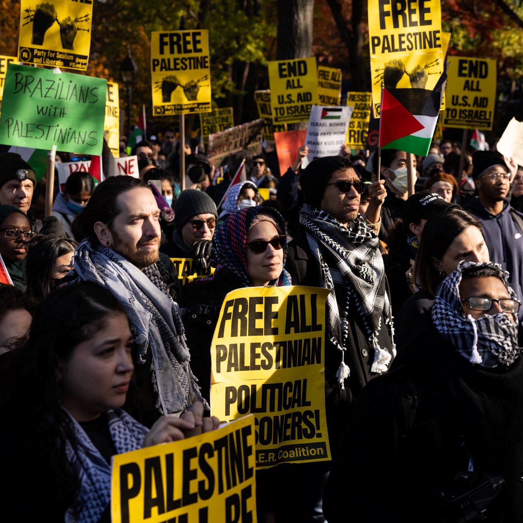 L'image montre une manifestation avec un grand nombre de personnes tenant des pancartes. Les pancartes portent des messages tels que "FREE PALESTINE" et "FREE ALL PALESTINIAN POLITICAL PRISONERS". Les participants sont vêtus de manière variée, certains portent des écharpes ou des keffiehs. On peut voir des drapeaux palestiniens en arrière-plan. L'ambiance semble être énergique et engagée, reflétant un fort sentiment de solidarité et de revendication pour les droits des Palestiniens.