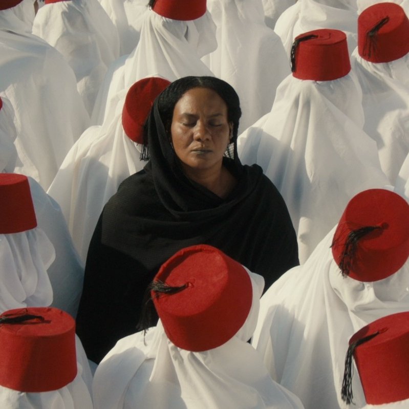L'image montre un groupe de personnes vêtues de longues robes blanches, portant des fez rouges. Au centre, une femme en noir se tient les yeux fermés, contrastant avec l'uniformité des autres. Cette composition crée une atmosphère de méditation ou de réflexion, avec un fort accent sur la spiritualité et la communauté. Les couleurs et les vêtements évoquent une tradition culturelle riche.