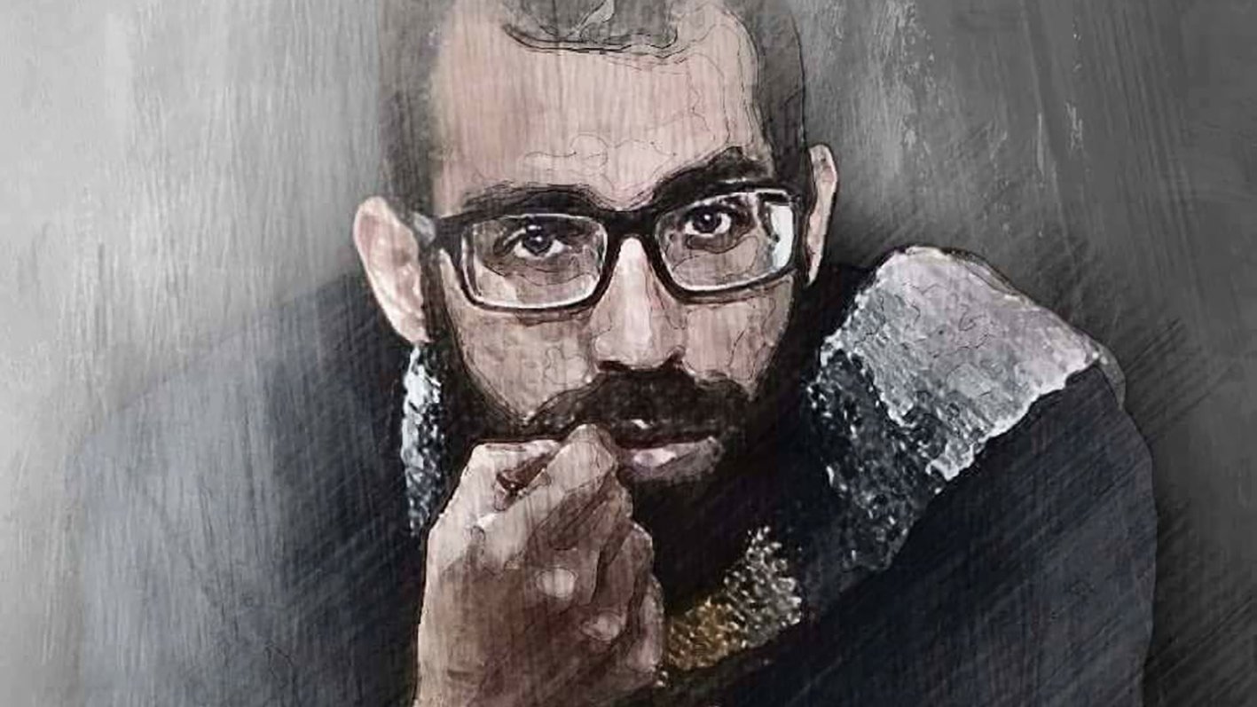 L'image montre un homme avec des lunettes, au regard intense, qui semble réfléchissant. Il a une barbe et porte un vêtement sombre avec des détails clairs. Le fond est flou et évoque une ambiance artistique ou mélancolique. Les traits de son visage sont bien définis, ce qui attire l'attention sur son expression.