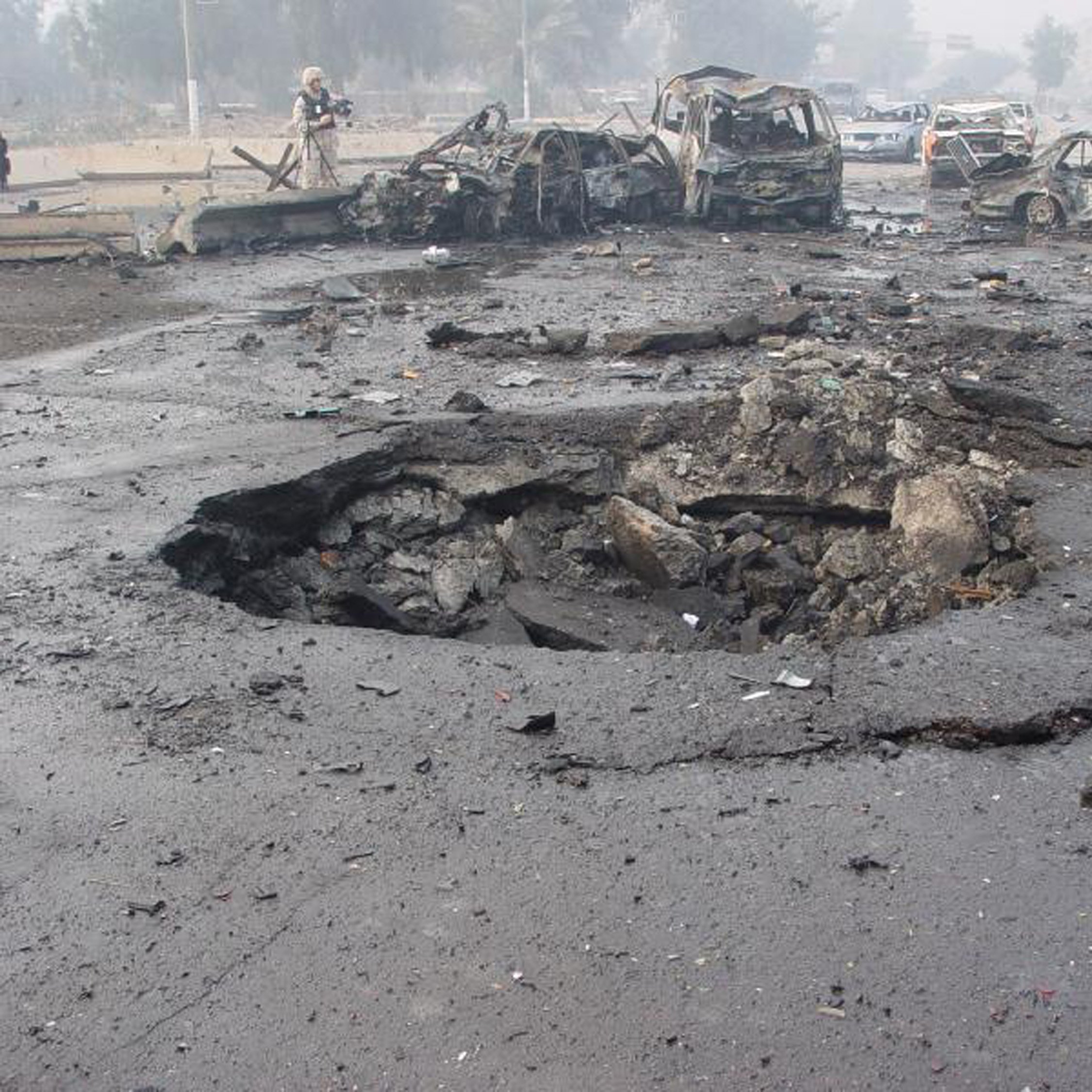 L'image montre un site de dévastation après une explosion. On peut voir plusieurs voitures endommagées et partiellement détruites, gisant sur le sol. Au centre, un cratère profond et large est présent dans le bitume, indiquant la force de l'explosion. L'environnement semble chaotique avec des débris éparpillés et une atmosphère troublée, probablement à cause de fumée ou de poussière. Cette scène évoque une situation d'urgence ou un acte de violence, reflétant des dommages importants.