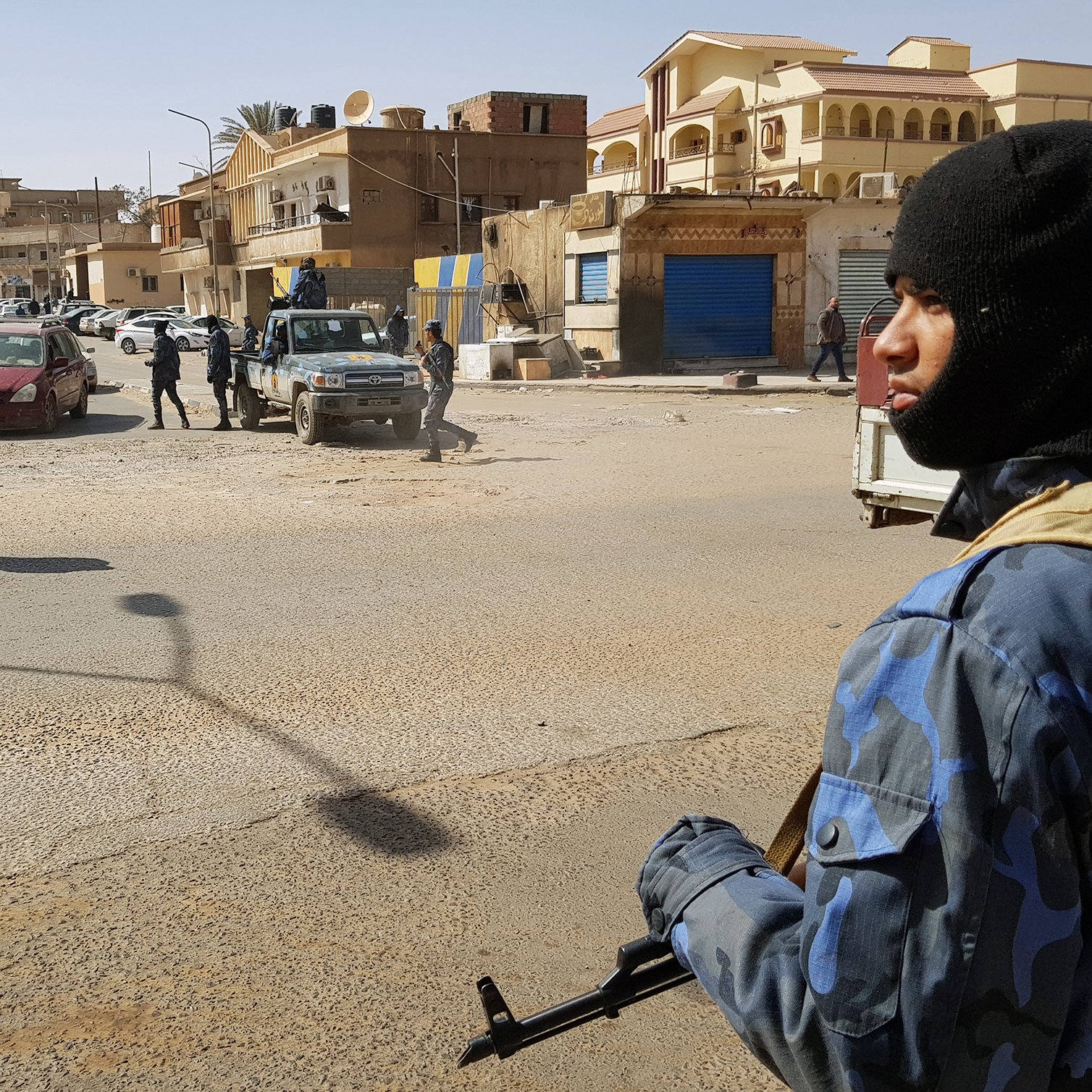 تظهر الصورة مشهداً في منطقة حضرية، حيث يوجد عدد من رجال الأمن يرتدون زيًّا رسميًّا وقبعات سُوداء. أحدهم يحمل سلاحًا ويقف في المقدمة، بينما يتواجد آخرون في الخلفية. تشهد المنطقة حركة مرورية، مع وجود سيارات متوقفة في الشارع. المباني المحيطة تظهر بألوان متنوعة، وبعضها يبدو أنه مغلق. تعكس الصورة أجواءً من اليقظة والحذر الأمني.