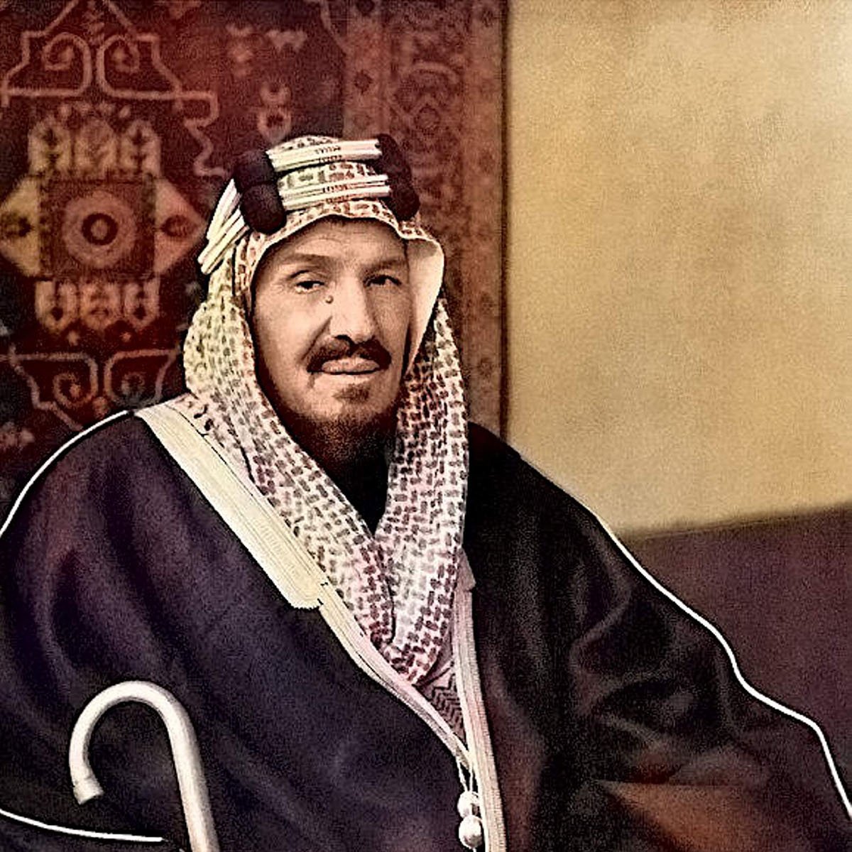 L'image montre un homme assis, portant un habit traditionnel. Il est vêtu d'une thobe sombre et d'un ghutrah, un couvre-chef caractéristique de la culture arabe. Il tient une canne et affiche une expression calme et posée. L'arrière-plan présente un motif décoratif qui renforce le contexte culturel de la scène. Les couleurs de l'image sont enrichies, donnant vie aux vêtements et à l'environnement.