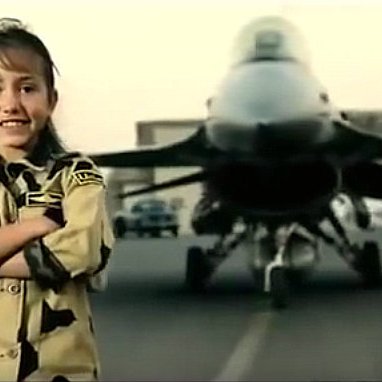 الصورة تظهر فتاة صغيرة تبتسم وتقف بفخر أمام طائرة مقاتلة. ترتدي زيًا عسكريًا يشبه الزي الذي يرتديه الجنود، ويبدو أنها تظهر الثقة والاعتزاز. خلفها، توجد الطائرة، مما يعطي انطباعًا عن القوة والجرأة. الأجواء في الصورة تبدو عسكرية، مع خلفية ترابية وألوان تدل على بيئة القاعدة العسكرية.