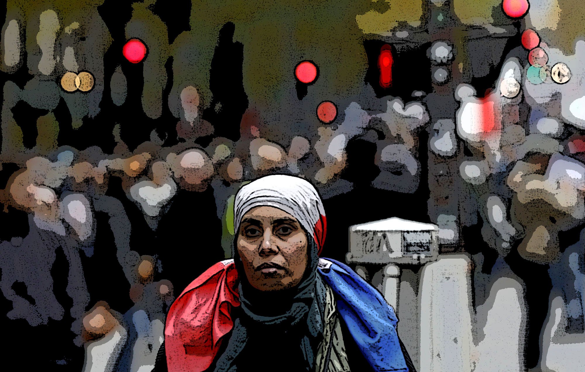 The image features a woman in the foreground wearing a white headscarf and a scarf that appears to have the colors of the French flag draped around her shoulders. She has a serious expression as she looks directly at the camera. The background is somewhat blurred and depicts a bustling crowd, suggesting a lively or busy environment, possibly a city street. The overall composition conveys a sense of solidarity or national identity amid a dynamic setting.