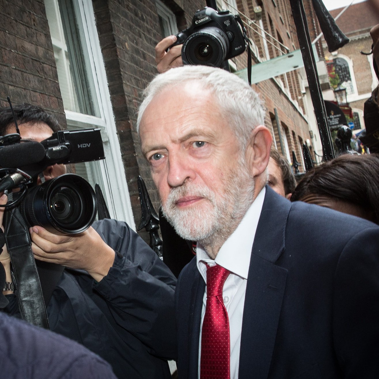 The image shows a man with gray hair and a beard, dressed in a dark suit and a red tie, walking through a crowd. He appears to be in a busy outdoor setting, surrounded by photographers and cameras capturing the moment. The buildings in the background suggest an urban environment. The overall atmosphere seems to convey a sense of public attention and media interest.