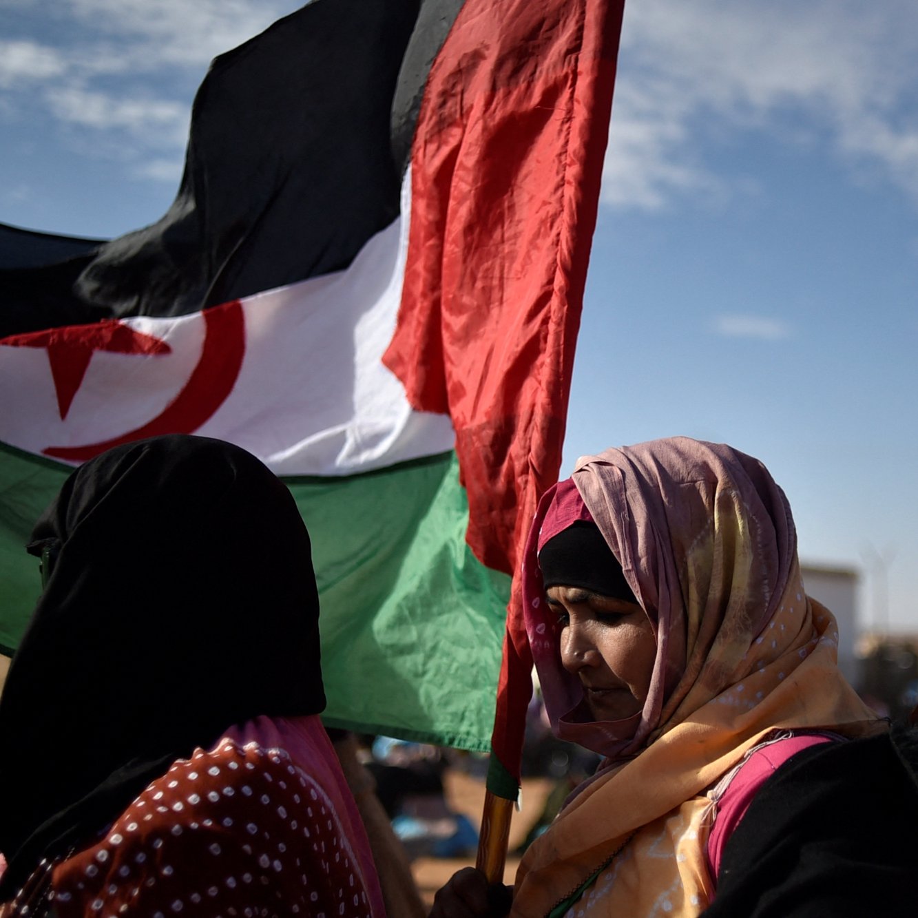 Des femmes portant une écharpe colorée et une grande bannière. Ciel dégagé en arrière-plan.