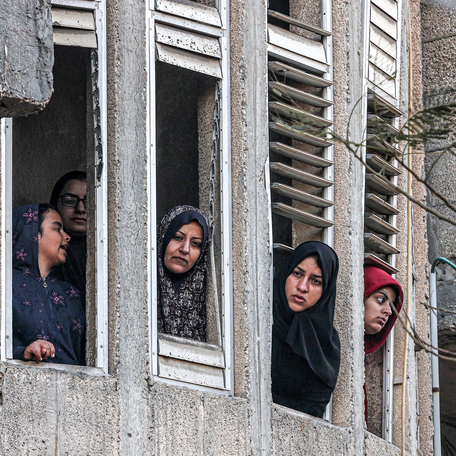 The image shows several women looking out of narrow windows in what appears to be a multi-story concrete building. The women are wearing headscarves and appear to be observing something outside with serious or concerned expressions. The building's exterior is worn and weathered, with visible signs of age and damage. The women are closely peering through the open windows, suggesting that they are trying to get a better view of something important happening outside. The overall mood of the image seems tense or somber.
