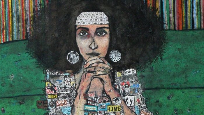 L'image représente une femme assise sur un canapé vert. Elle a de grands cheveux crépus et porte un bandeau blanc. Ses mains sont jointes sous son menton, et elle affiche une expression pensive. Son corps est recouvert de collage, composé de divers autocollants et affiches, représentant des mots et des images liées à des thèmes comme les droits, la révolution, et la maison. En arrière-plan, on observe des bandes colorées, ajoutant une ambiance vibrante à la scène.