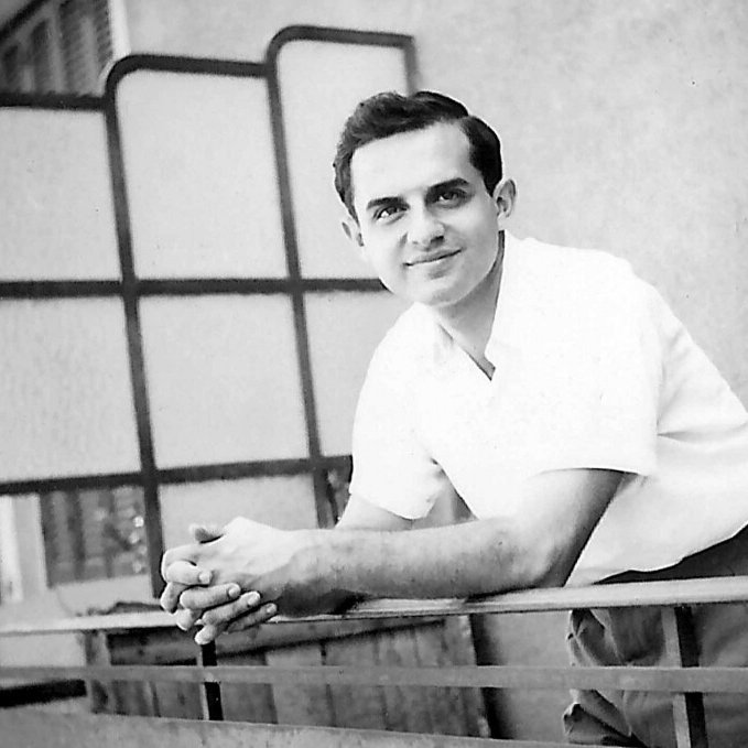 در این تصویر، مردی جوان به نظر می‌رسد که در حال لبخند زدن است. او با آستین‌های کوتاه و پیراهن سفیدی لباس پوشیده و در بالکن یا جایی مشابه ایستاده است. دستانش را بر روی نرده‌های بالکن گذاشته و به دوربین نگاه می‌کند. پس‌زمینه تصویر شامل دیواری ساده و پنجره‌ای با شیشه‌های مشبک است. جو تصویر دوستانه و صمیمانه به نظر می‌رسد.