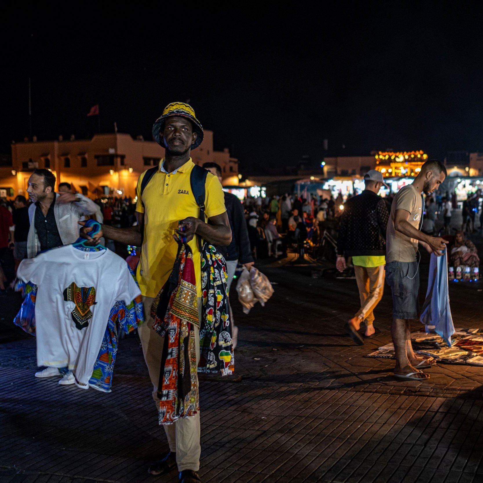 L'image montre un marché nocturne animé, probablement dans un endroit comme Marrakech. On peut voir des gens se déplacer dans une ambiance vibrante. Au premier plan, un homme porte un t-shirt jaune et tient plusieurs vêtements traditionnels colorés. En arrière-plan, d'autres personnes flânent et explorent les étals, tandis que des lumières scintillantes créent une atmosphère festive. Des tapis et d'autres articles sont visibles au sol, ajoutant à l'attrait du marché.