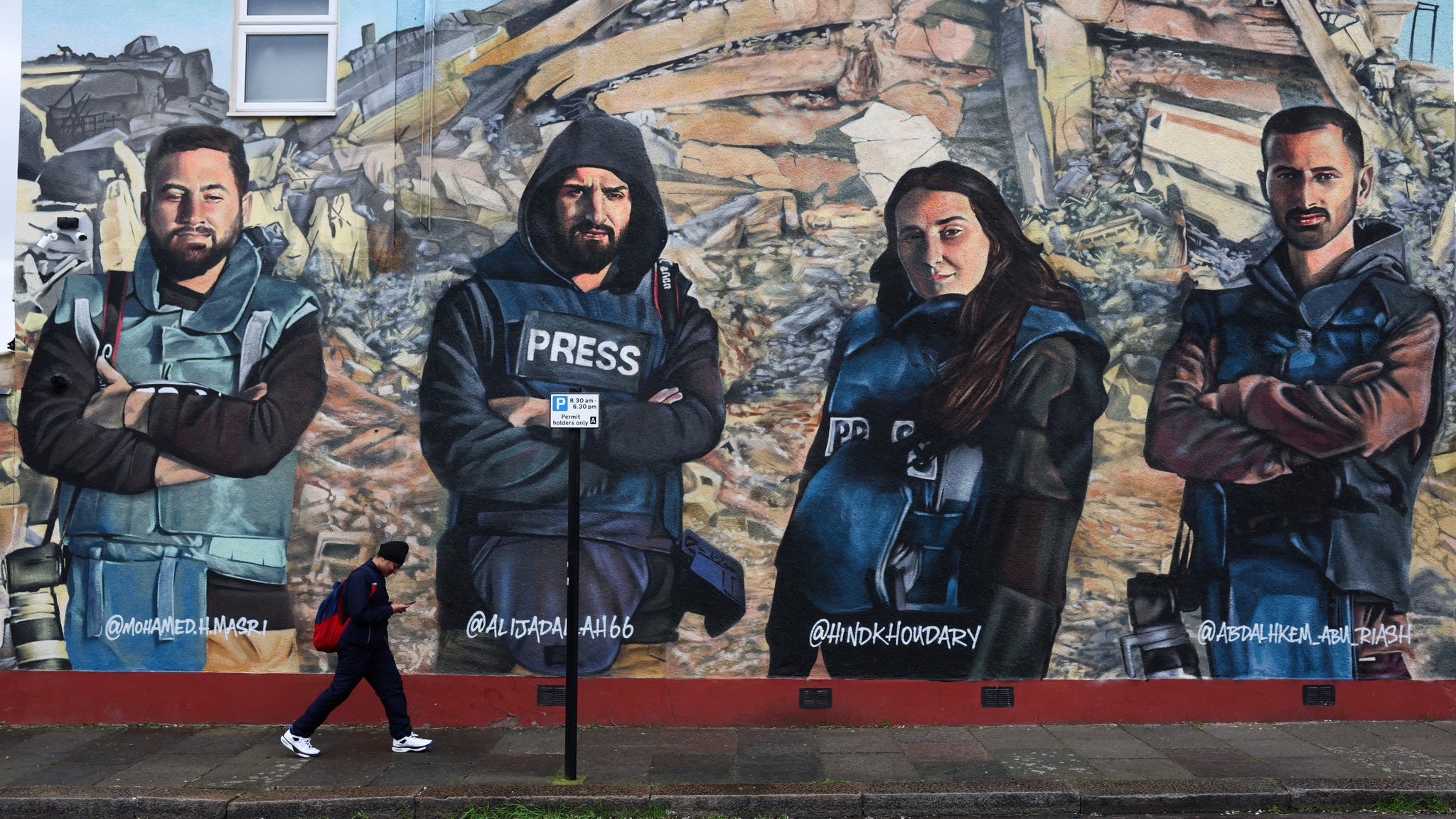 L'immagine mostra un murale che ritrae quattro giornalisti con abbigliamento di protezione e giubbotti con la scritta "PRESS". Sono posizionati in modo prominente davanti a un contesto di distruzione, simile a un'area colpita da un terremoto. Una persona sta camminando lungo il marciapiede di fronte al murale. I volti dei giornalisti esprimono determinazione e serietà, sottolineando il loro lavoro e l'importanza della libertà di stampa.
