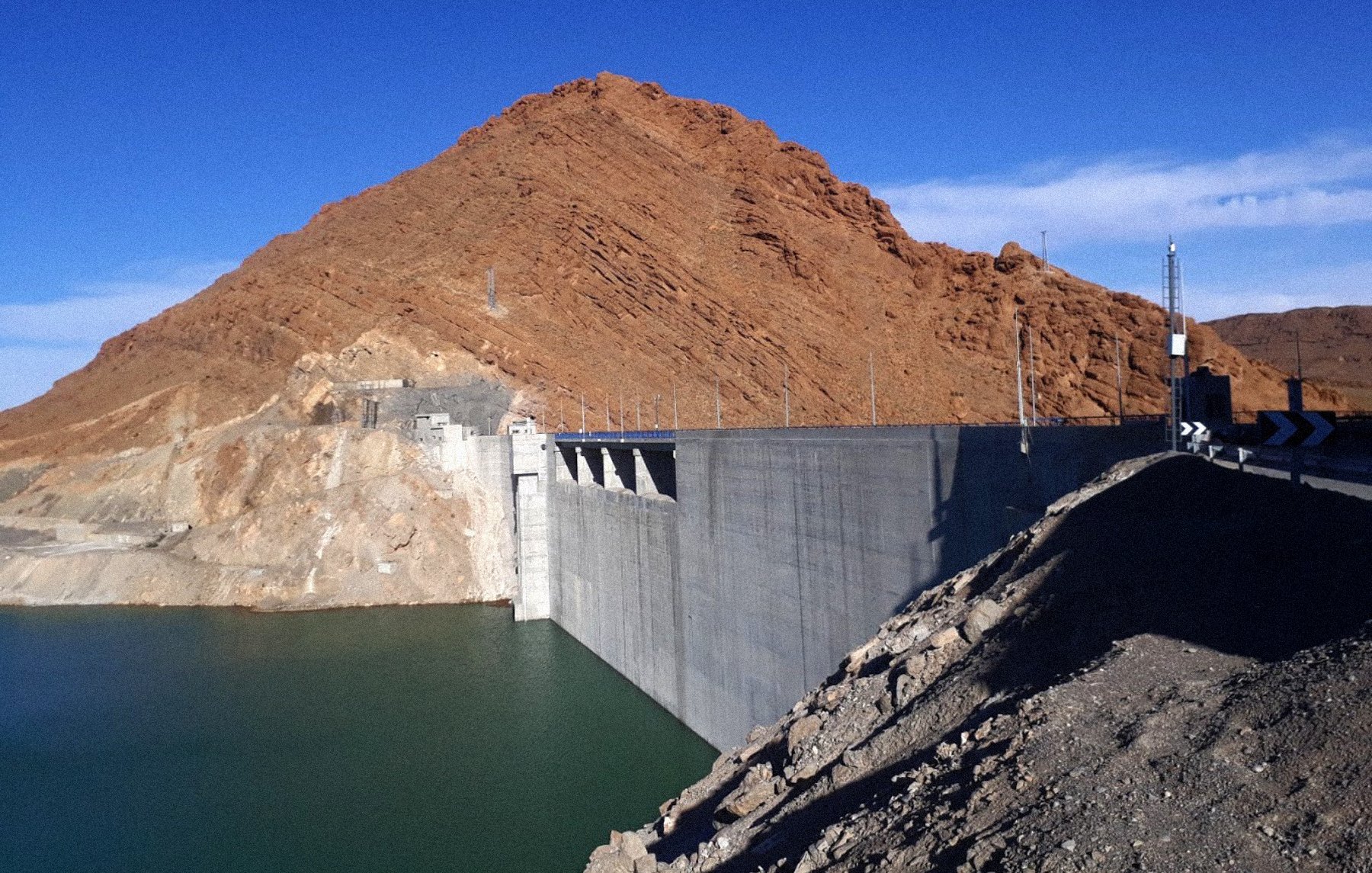 L'image montre un barrage construit sur un cours d'eau, avec une grande structure en béton qui s'étend le long de la rive. À l'arrière-plan, on voit une montagne aride, aux pentes abruptes et aux teintes brunes. Le ciel est dégagé, d'un bleu vif, et l'eau du réservoir est calme et d'une teinte verte-bleue. Le paysage semble sec et stérile, typique des régions désertiques.