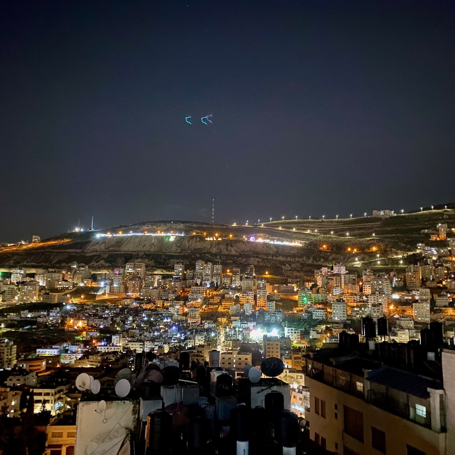 L'immagine mostra una vista notturna di una città, con luci brillanti che illuminano gli edifici. Sullo sfondo si possono vedere colline, anch'esse parzialmente illuminate. Gli edifici sono disposti su vari livelli, creando un effetto panoramico. La scena trasmette un'atmosfera vivace e dinamica, tipica di un centro urbano notturno. La presenza di antenne e strutture sul tetto di alcuni edifici suggerisce un'area residenziale o commerciale.