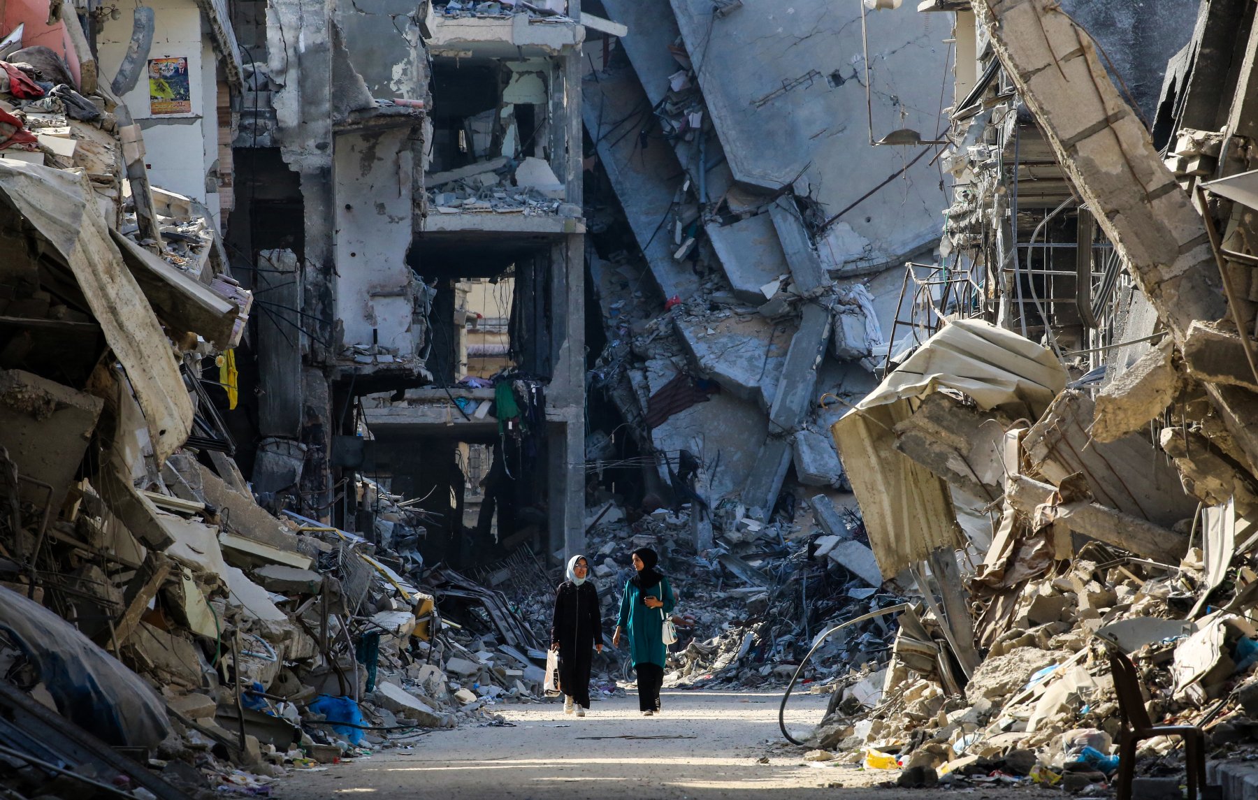 تصویر نشان‌دهنده خیابانی است که در میان ویرانه‌های ساختمان‌ها قرار دارد. دیوارهای بتنی و تخریب‌شده در دو طرف خیابان وجود دارند و debris و زباله‌ها در زمین پخش شده‌اند. دو زن در حال راه رفتن در این خیابان هستند و به نظر می‌رسد در جستجوی امنیت یا کمک هستند. نور خورشید به آرامی از میان ویرانه‌ها عبور می‌کند و سایه‌های طولانی‌ای بر زمین می‌افکند. حس و حال تصویر، غم و ویرانی را به نمایش می‌گذارد.