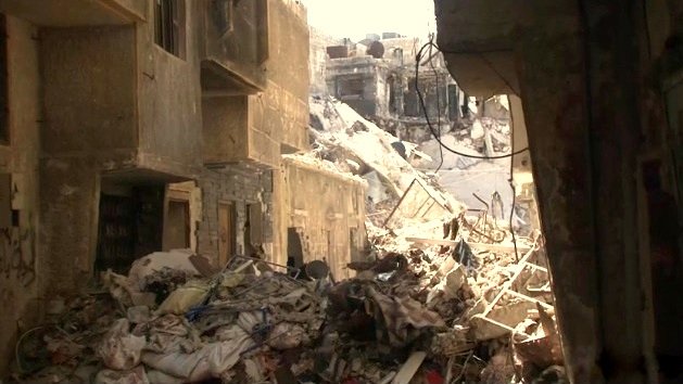 L'image montre une scène de destruction, probablement dans une zone urbaine touchée par un conflit ou un désastre. Des bâtiments sont en ruines, avec des murs effondrés et des débris éparpillés sur le sol. On peut voir des tas de gravats et de déchets, ce qui suggère que la zone a été abandonnée ou négligée. La lumière du soleil pénètre à travers les décombres, créant un contraste entre l'ombre et la lumière. L'atmosphère évoque la désolation et la tristesse.