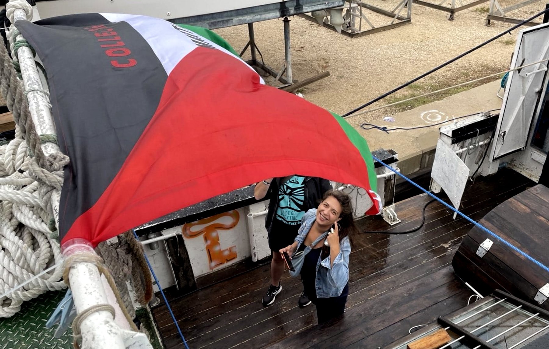 Nell'immagine si possono vedere due persone che stanno sistemando una grande bandiera, verosimilmente di un paese, su una barca o una piattaforma di legno. Una delle persone sembra sorridere e fare un gesto di saluto, mentre l'altra è occupata a sistemare la bandiera. La bandiera presenta i colori verde, bianco e rosso, tipici di alcune bandiere nazionali. Lo sfondo è composto da strutture marine e un'atmosfera marittima.