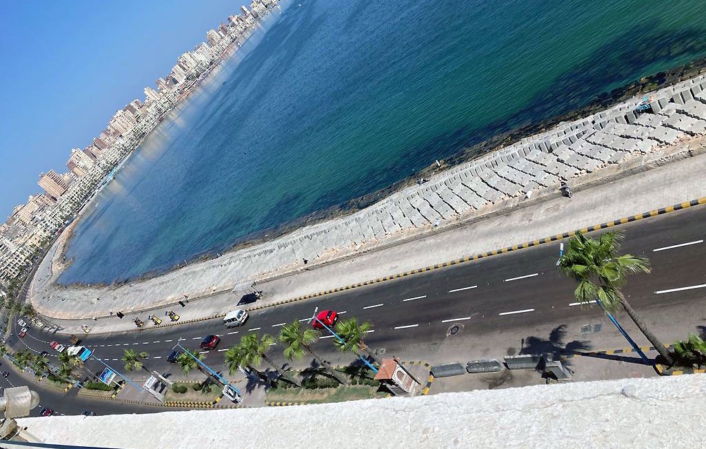 L'image montre une vue panoramique d'une côte en bord de mer. On aperçoit une route qui longe la plage, avec des palmiers sur le côté. L'eau se présente dans des teintes de bleu et de vert, et on peut voir des bâtiments en fond, probablement la ville ou des complexes résidentiels. La lumière du soleil illumine la scène, donnant une atmosphère agréable et ensoleillée.