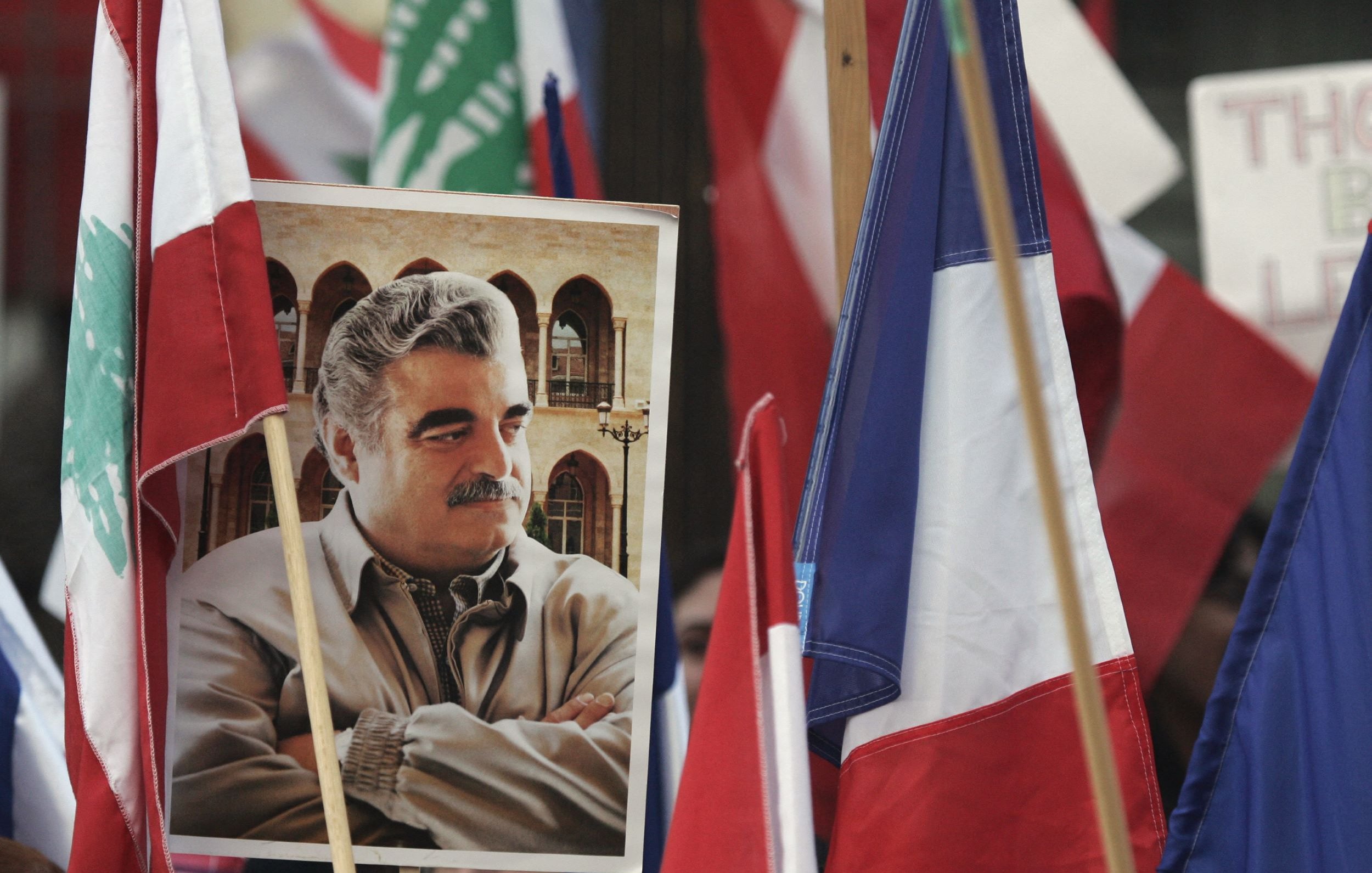 تظهر في هذه الصورة مجموعة من الأعلام، بما في ذلك العلم اللبناني والعلم الفرنسي. في وسط الصورة، هناك صورة كبيرة لرجل يبدو أنه شخصية معروفة، حيث يظهر واقفًا مع ذراعيه متقاطعتين. الخلفية تضم مجموعة من الأشخاص الذين يحملون الأعلام، مما يدل على أن هذه الصورة قد التقطت خلال فعالية أو تجمع يمثل قضية معينة.