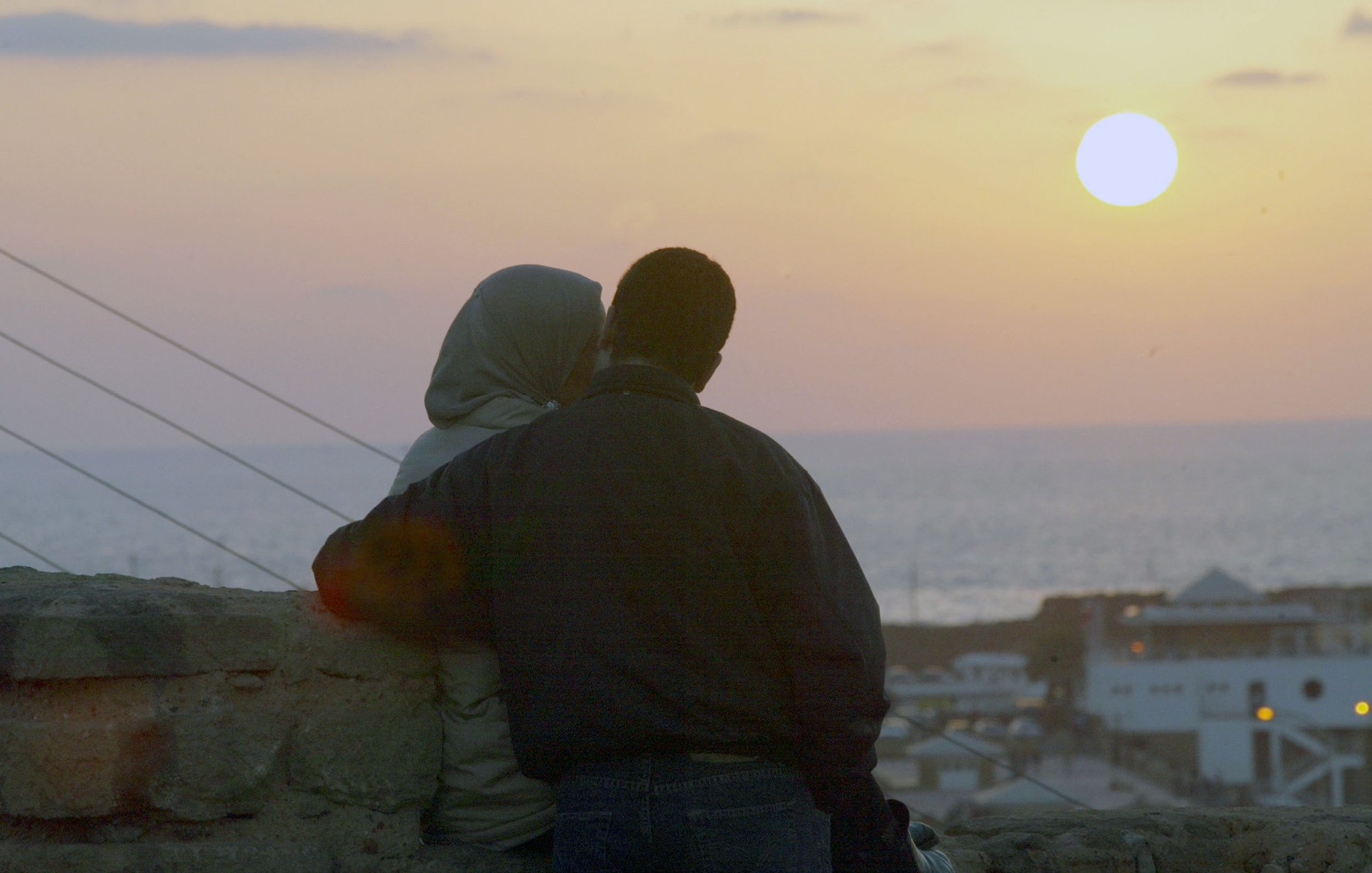 L'image montre un couple assis côte à côte, observant le coucher de soleil. L'un des membres du couple porte un hijab, tandis que l'autre est vêtu d'une veste sombre. Ils semblent se rapprocher l'un de l'autre, créant une atmosphère intime. En arrière-plan, on peut voir la mer, et le ciel est teinté de couleurs douces, avec le soleil se couchant à l'horizon. Les bâtiments sont légèrement visibles en bas, ajoutant à la scène romantique.