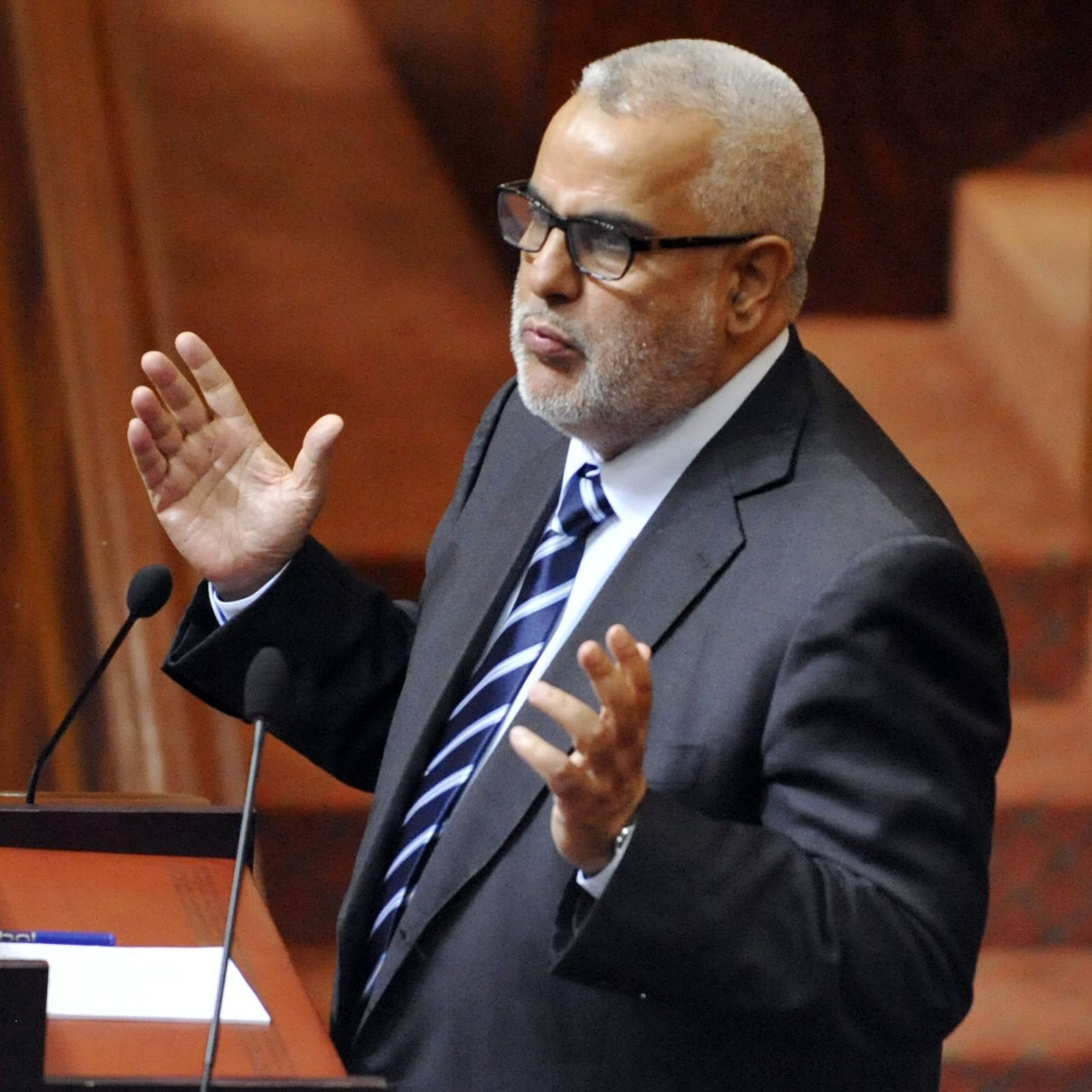 L'image montre un homme debout devant un pupitre, probablement en train de s'exprimer lors d'une réunion ou d'une séance législative. Il porte un costume sombre avec une chemise et une cravate à rayures. Son attitude et ses gestes suggèrent qu'il est engagé dans un discours ou une argumentation. L'environnement semble être celui d'une assemblée, avec des éléments de décor en bois et des sièges visibles en arrière-plan.