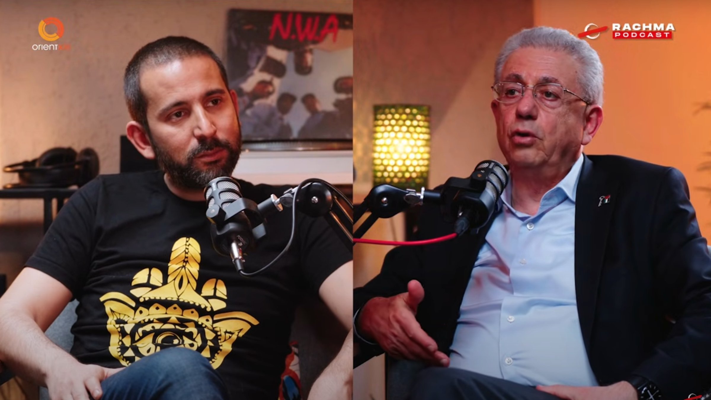 في الصورة، يظهر شخصان يجلسان معاً، يتحدثان، وأمام كل منهما ميكروفون.