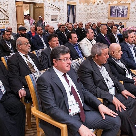 L'image montre un groupe de personnes assises, probablement lors d'une réunion ou d'une conférence. Ils semblent être des hommes d'affaires ou des responsables, vêtus de costumes formels. L'arrière-plan est orné de décorations élégantes, typiques d'une salle de conférence dans un cadre sophistiqué. Les participants regardent attentivement, suggérant un moment de discussion ou de présentation importante.