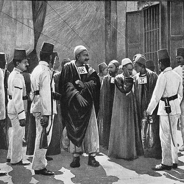 L'image présente un groupe de personnes en tenue traditionnelle. Au centre, un homme en robe longue semble diriger la scène, tandis que d'autres, habillés en uniformes ou en costumes distinctifs, l'entourent. Certains des individus portent des chapeaux spécifiques, comme des fez. Ils semblent engagés dans une conversation ou une activité, suggérant une interaction communautaire ou une réunion formelle. L'arrière-plan indique un environnement urbain, peut-être une scène historique. La composition dégage une atmosphère d'autorité et de respect.