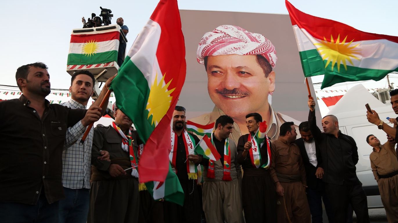 L'image montre un groupe de personnes tenant des drapeaux kurdes, célébrant devant une grande affiche d'une personnalité kurde. Les participants portent des vêtements traditionnels et semblent joyeux, suggérant une ambiance festive. L'affiche derrière eux représente un homme souriant, probablement un leader ou une figure importante dans la culture kurde. Les décorations et les drapeaux créent une atmosphère vibrante et patriotique.