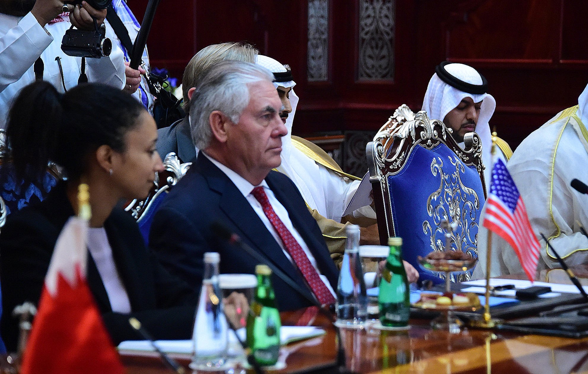 L'image présente une réunion officielle, où l'on peut voir plusieurs personnes assises autour d'une grande table. À gauche, une femme est accoudée à la table, tandis qu'à droite, un homme en costume, probablement un diplomate, semble écouter attentivement. Au centre, il y a un drapeau américain, et derrière, d'autres participants portant des vêtements traditionnels peuvent être aperçus. L'environnement semble formel, avec une décoration riche et des bouteilles d'eau sur la table.