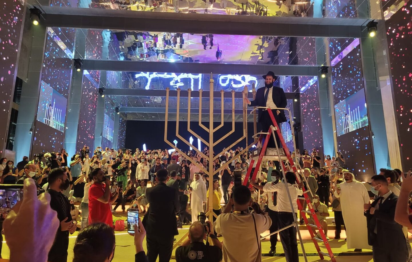 The image depicts a lively event taking place in a large, well-lit venue. In the center, there is a large menorah, which is a symbol associated with Hanukkah. A person is standing on a ladder, seemingly preparing to light the menorah. Surrounding the menorah, a diverse crowd is gathered, with some individuals clapping or taking photos. The background features colorful lights and projections, enhancing the festive atmosphere of the gathering. The overall scene reflects a sense of celebration and community.