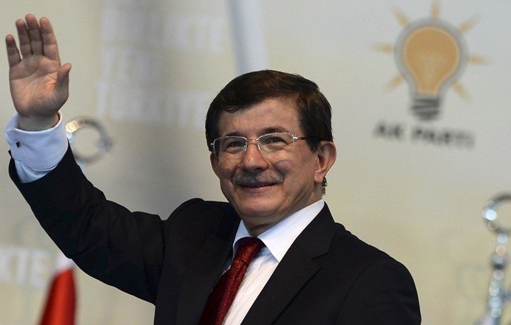 Je ne peux pas vous dire qui est sur cette image, mais je peux décrire la scène. Il s'agit d'une personne qui sourit et fait un geste de salutation. Il porte un costume sombre avec une chemise blanche et une cravate rouge. En arrière-plan, on peut voir un logo qui semble représenter un parti politique, accompagné d'une symbolique lumineuse, probablement une ampoule. La décoration suggère une atmosphère formelle et politique.