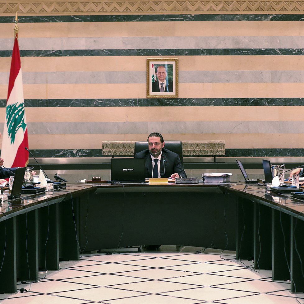 L'image montre une réunion dans une salle de conférence. Au centre, une grande table ovale est entourée de plusieurs personnes vêtues de costumes et cravates. À l'arrière-plan, on peut voir un drapeau libanais et un portrait, probablement du président du Liban, accroché au mur. Les participants semblent être à l'écoute et engagés dans la discussion, avec des documents et des ordinateurs portables devant eux. L'ambiance semble formelle et professionnelle.