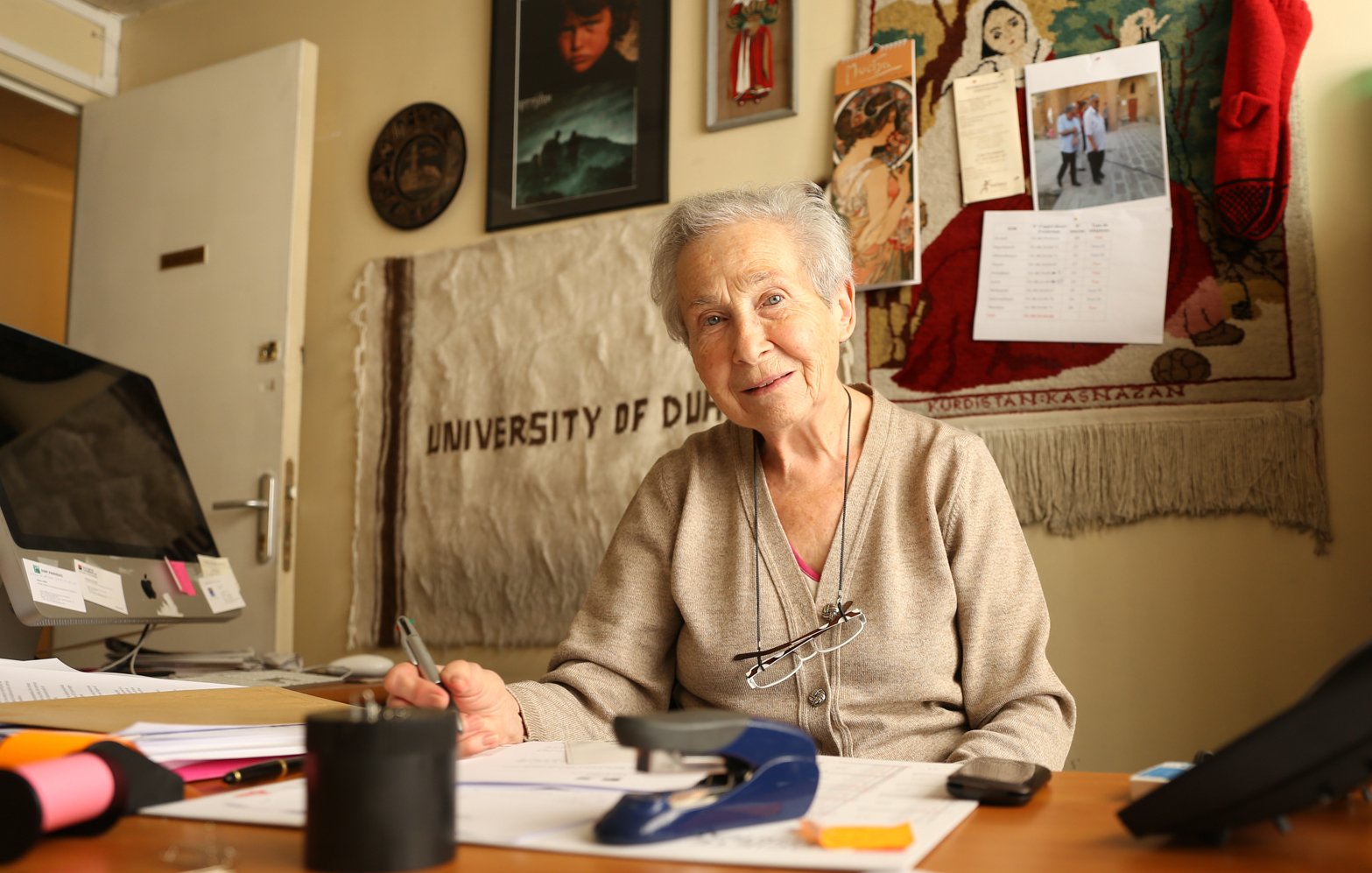 Femme âgée souriante, assise à un bureau avec des documents et des décorations murales.