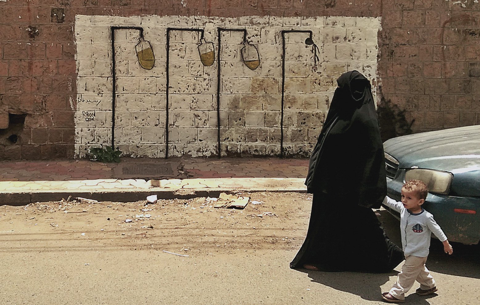 L'image montre une femme vêtue d'une tenue noire, probablement un hijab ou une burqa, marchant dans une rue. À ses côtés, un enfant tient sa main. En arrière-plan, on aperçoit un mur de béton avec un graffiti qui représente des pots de peinture suspendus, ajoutant une touche artistique à la scène. La route semble modeste avec des débris au sol, ce qui donne un aperçu de l'environnement urbain.