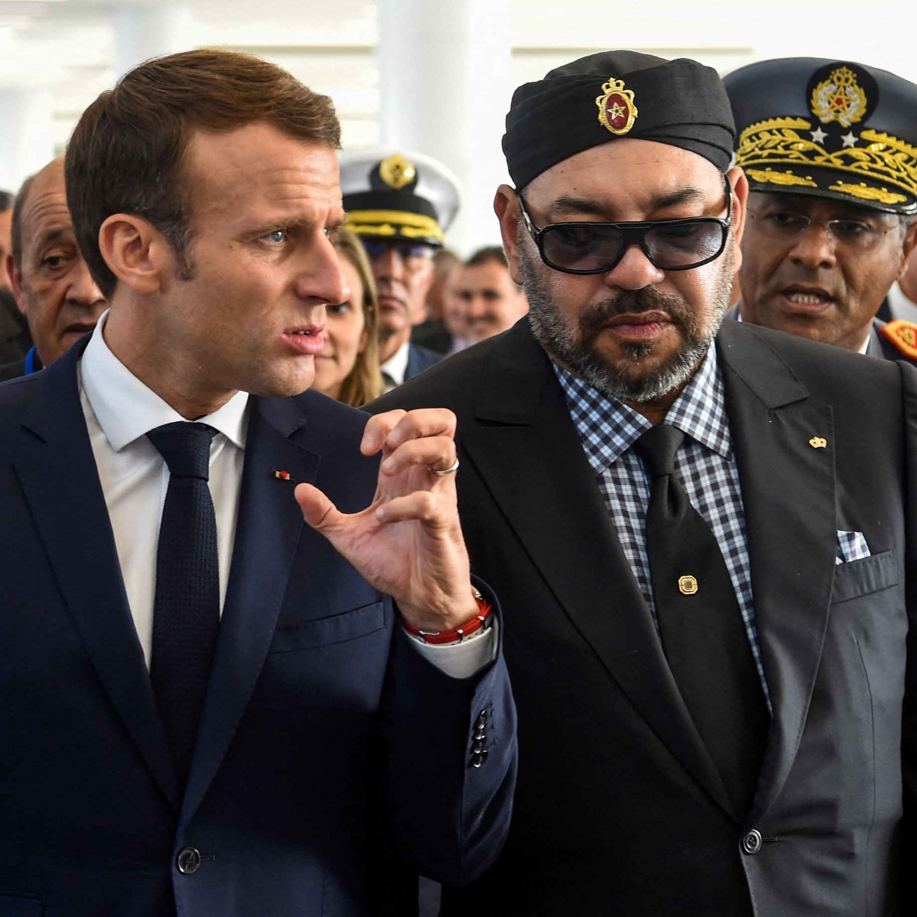 L'image montre deux hommes en discussion, entourés de plusieurs personnes. L'un porte un costume sombre et une cravate, tandis que l'autre est habillé d'une manière plus formelle, avec des lunettes de soleil et un turban. Ils semblent engagés dans une conversation sérieuse, et l'environnement semble être une cérémonie officielle ou un événement important. Les expressions sur leurs visages suggèrent qu'ils sont en train de partager des idées ou des informations.