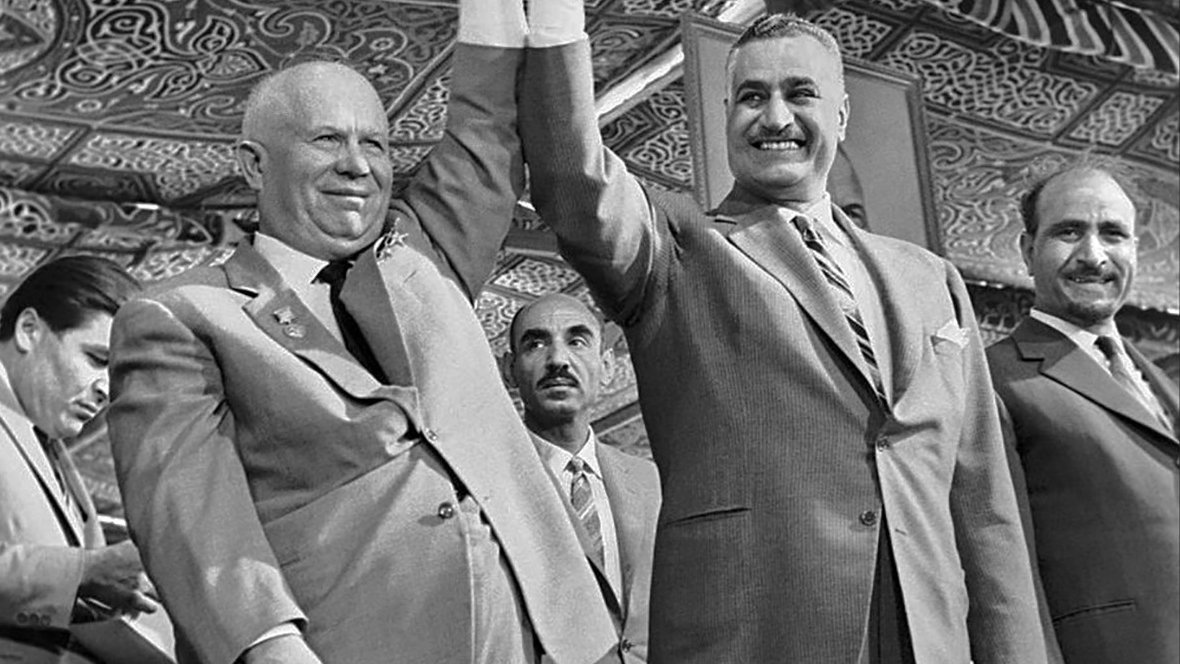 L'image montre deux hommes se tenant la main, souriant et levant les bras en signe de victoire ou de camaraderie. Ils sont entourés de plusieurs autres hommes en arrière-plan, qui semblent également participer à l'événement. Les tenues des protagonistes suggèrent un contexte formel ou officiel, avec des costumes élégants. Le décor en arrière-plan présente des motifs ornementaux, indiquant un événement culturel ou politique. L'ambiance générale semble être celle d'une célébration ou d'une réunion importante.