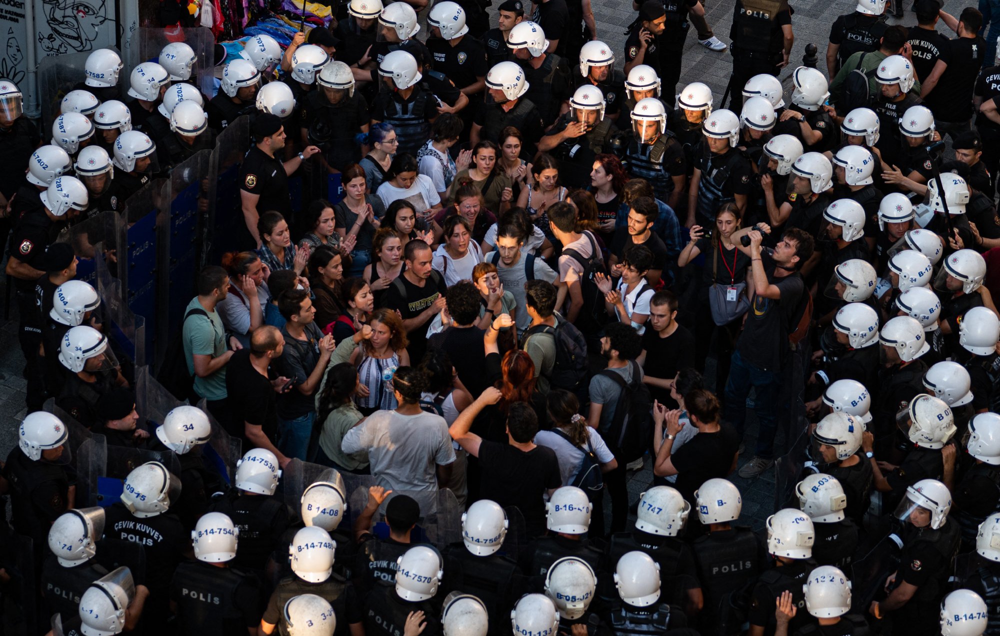در این تصویر، گروهی از مردم در حال تظاهرات یا تجمعی هستند که با نیروهای پلیس محاصره شده‌اند. افراد معترض در وسط تصویر قرار دارند و به نظر می‌رسد در حال گفت‌وگو یا گفتگو با پلیس هستند. نیروهای پلیس در اطراف آنها ایستاده‌اند و کلاه‌های مخصوصی به سر دارند. فضای تصویر نشان‌دهنده تنش و فعالیت اجتماعی است، با تجمع جمعیتی که به سمت نیروهای پلیس نگاه می‌کنند.