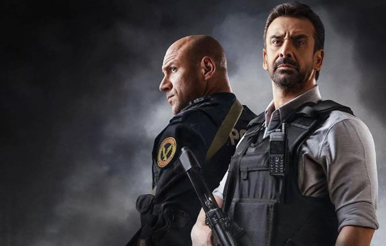 الصورة تظهر شخصين يرتديان زي الشرطة ويقفان معًا. أحدهما يبدو في وضعية أكثر تكاملًا، حيث يحمل سلاحًا ويعبر عن جدية معينة. الخلفية داكنة مما يعطي صورة درامية وينقل إحساسًا بالتوتر أو الإثارة. الشخصان يُظهران تعابير وجه تتسم بالتركيز، مما يوحي بأنهما في موقف يتطلب الحذر والانتباه.