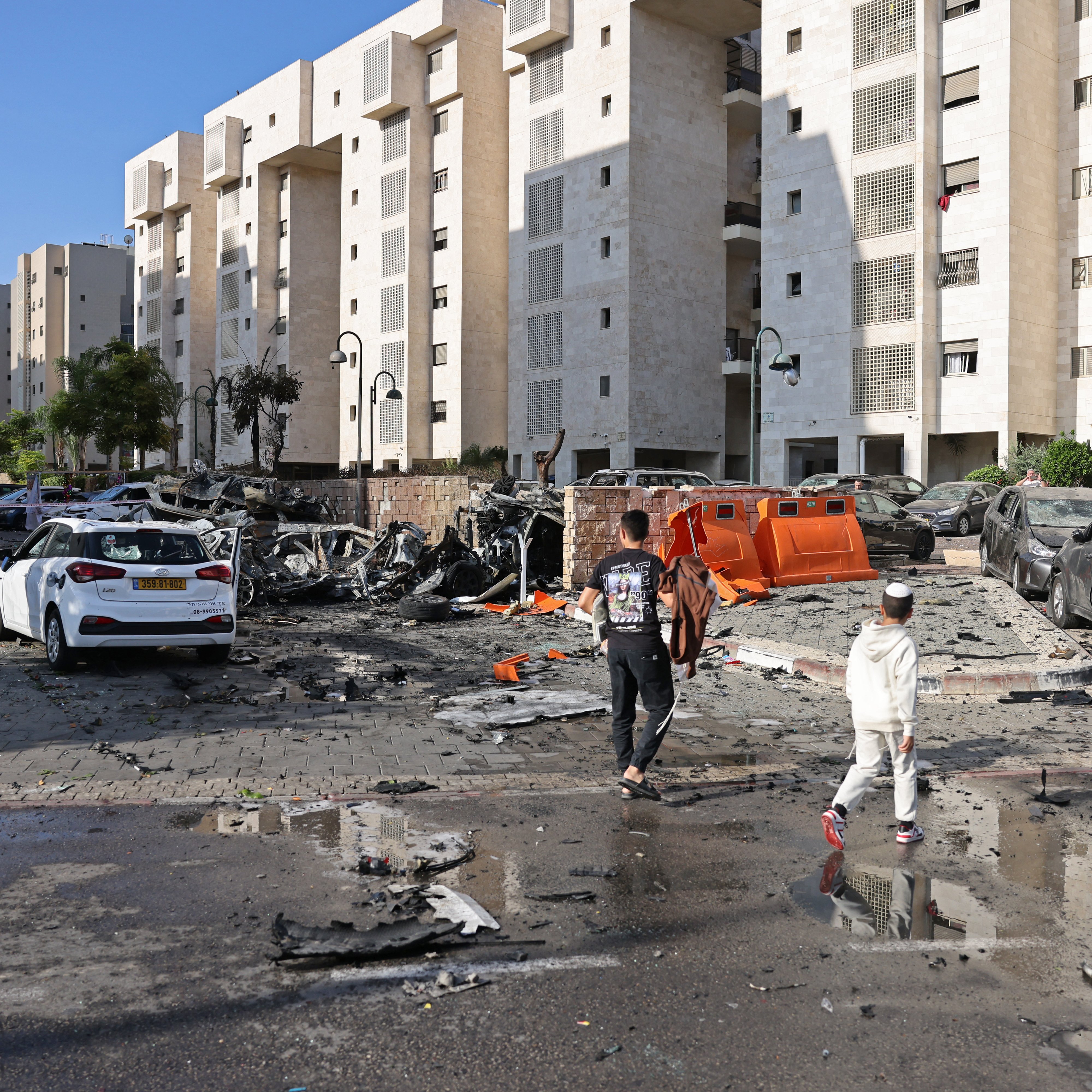 L'immagine mostra una scena di distruzione in un'area urbana. Si possono vedere edifici residenziali con facciate chiare, circondati da macerie e detriti. Alcune auto sono danneggiate, con pezzi sparsi sul terreno. Ci sono persone che camminano nella strada, apparentemente mentre osservano il danno. Il cielo è sereno, e ci sono anche pozzanghere d'acqua, suggesting che recente pioggia potrebbe essere rientrata nell'area. La scena trasmette un senso di caos e di recente violenza o attacco.