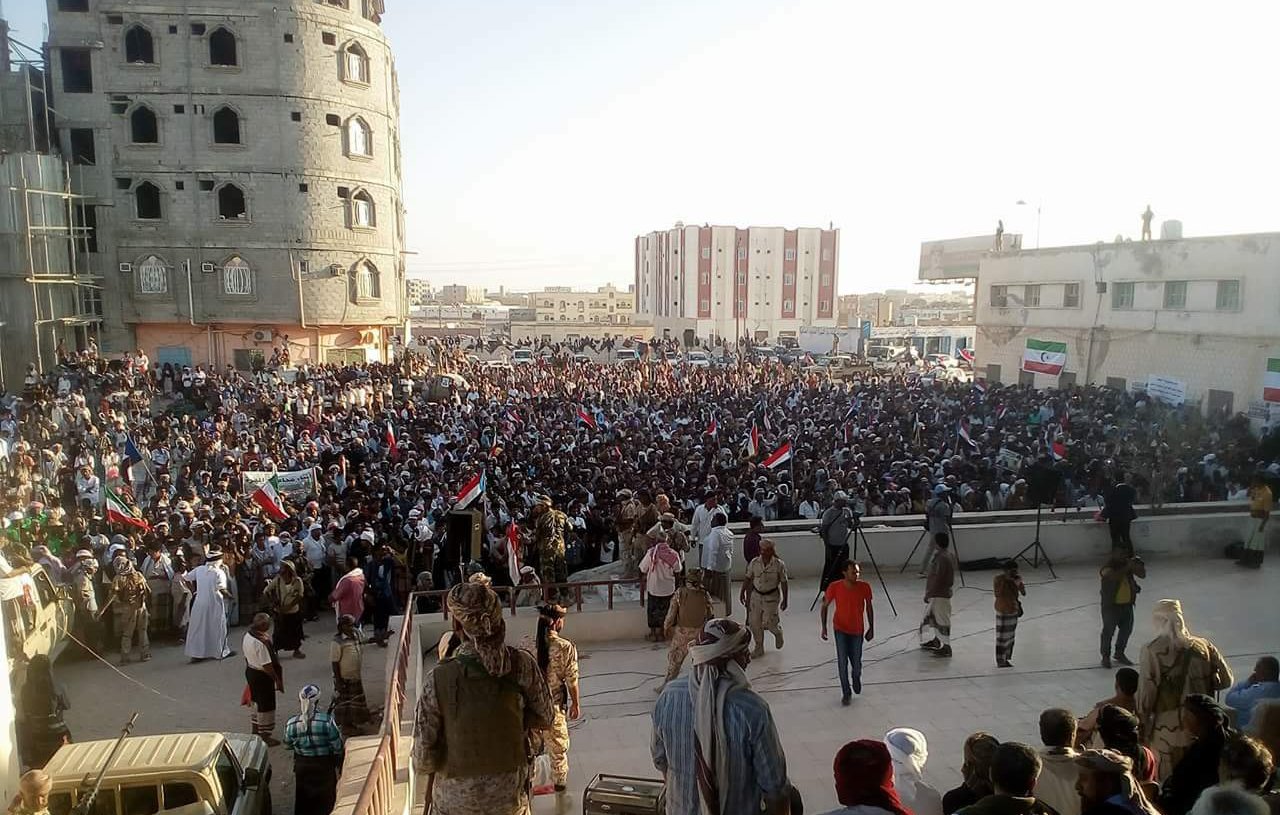 L'image montre une grande foule rassemblée lors d'une manifestation ou d'un rassemblement public. Les participants tiennent des banderoles et des drapeaux, indiquant un fort engagement politique ou social. En arrière-plan, on peut voir des bâtiments en construction ou rénovés, ainsi qu'une architecture variée. La scène semble se dérouler en plein air, probablement durant l'après-midi ou le début de la soirée, avec une lumière naturelle éclatante. L'atmosphère est dynamique, avec beaucoup de gens engagés dans une cause commune.
