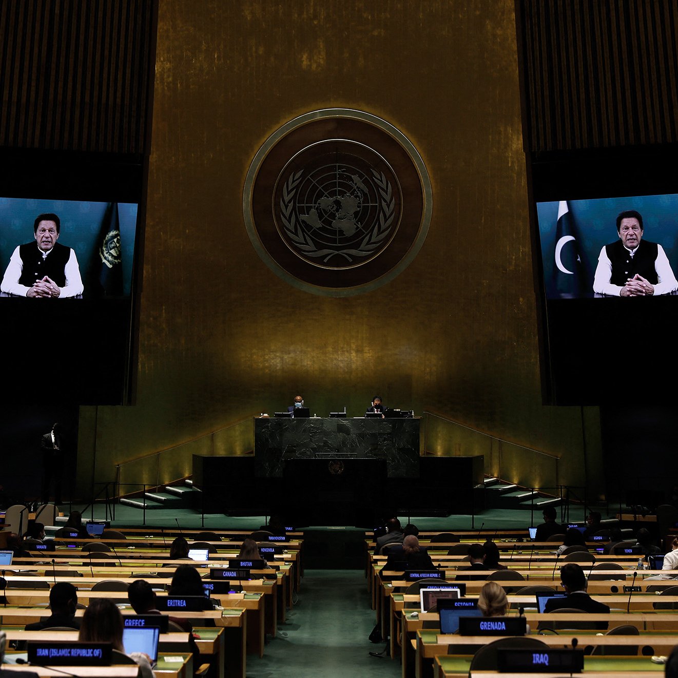 L'image montre une scène à l'Assemblée générale des Nations Unies. On peut voir des délégués assis à des bureaux, prêts à participer à la réunion. Un grand écran affiche une personne qui s'adresse à l'assemblée, tandis que d'autres officiels sont visibles sur la scène principale derrière le pupitre. L'arrière-plan est décoré avec l'emblème des Nations Unies, symbolisant l'importance de cet organe international. L'atmosphère est formelle et solennelle.