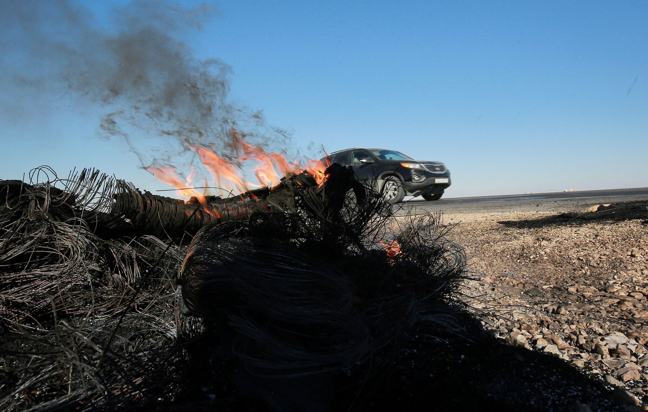 L'image montre un véhicule qui roule rapidement le long d'une route, tandis qu'un feu brûle sur le bord de la route. De la fumée s'élève du feu, qui semble être alimenté par des matériaux tels que des câbles ou des filets. Le ciel est dégagé avec une couleur bleue, et le paysage environnant est aride. On peut voir des débris brûlés au premier plan, ce qui suggère une scène de destruction ou d'accident.