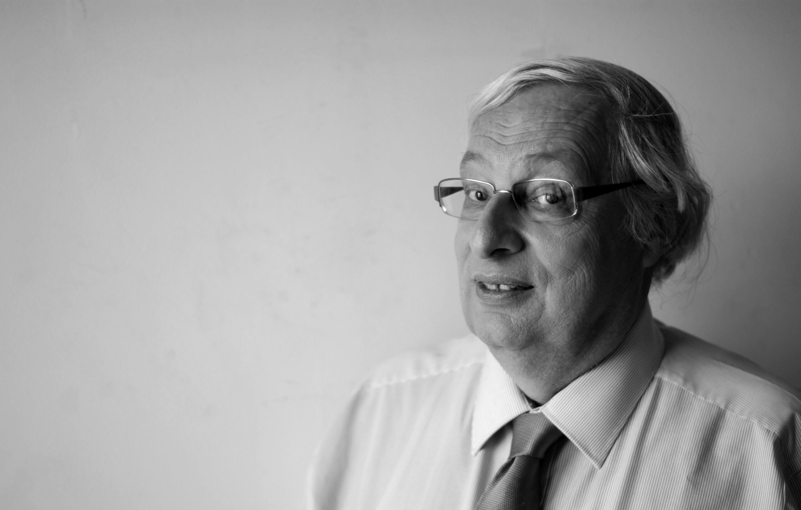 L'image montre un homme d'âge moyen avec des cheveux gris et des lunettes. Il porte une chemise claire avec une cravate. Il regarde légèrement sur le côté avec un sourire, le fond étant neutre et clair. L'image est en noir et blanc, ce qui lui donne un aspect classique et intemporel.