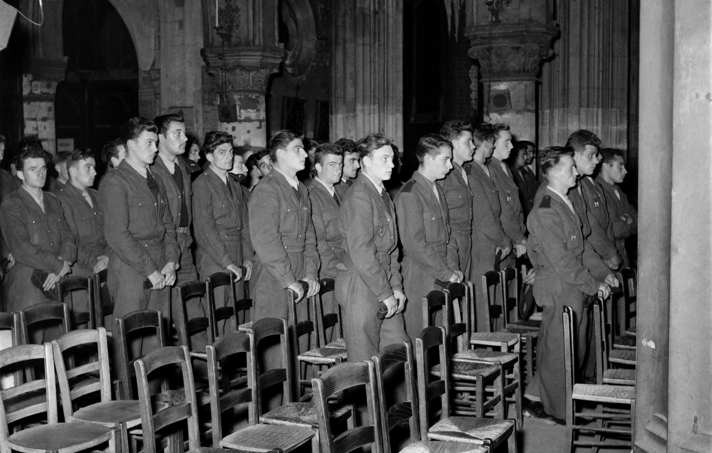 L'image montre un groupe de jeunes hommes en uniforme, probablement des soldats, se tenant debout en rang dans une église ou un bâtiment religieux. Ils semblent attentifs et concentrés, se tenant droit, dans un environnement décoré d'éléments architecturaux gothiques. Des chaises en bois sont visibles devant eux, et l'éclairage met en valeur l'atmosphère solennelle du lieu.