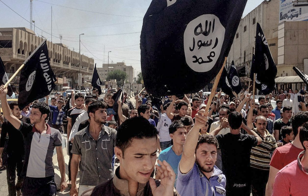 L'image montre une foule de personnes rassemblées dans une rue, tenant des drapeaux noirs emblématiques. Les manifestants semblent mobilisés et expriment une forte émotion. L'environnement urbain autour d'eux montre des bâtiments et une rue animée, suggérant un événement significatif ou une manifestation.